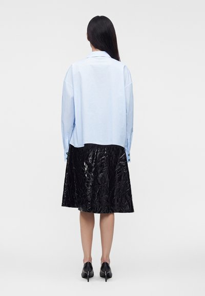 Femme aux cheveux longs et foncés portant une chemise bleu clair à manches longues, une jupe midi noire texturée et des chaussures noires à talons hauts, de dos.