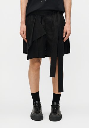 Homme portant un short asymétrique noir avec de longues cordelettes, des chaussettes noires et des baskets noires, visible de la mi-cuisse aux pieds.