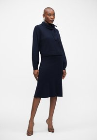 Μπλε μαρινάρια πλεκτό φόρεμα με ταιριαστό κοντό πουλόβερ που διαθέτει διπλωμένο γιακά και μακριά μανίκια, φτιαγμένο από μαλακό ύφασμα με λεία υφή.