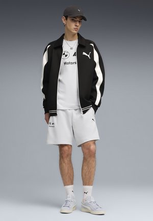 Młody mężczyzna ubrany w czarno-białą kurtkę Puma, białą koszulkę BMW Motorsport i szorty, szare buty Puma oraz czapkę, stojący na tle szarego tła.