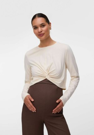 Frau mit dunklen Haaren in einem cremefarbenen, verdrehten Langarm-Top und braunen, hoch taillierten Hosen hält sanft ihren Bauch und lächelt sanft.