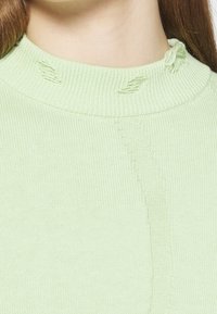 Ljusgrön stickad tröja med rund hals, med slitna detaljer längs halsringningen och en mjuk textur.