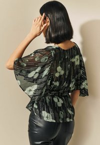 Blouse noire et verte à motifs avec tissu transparent, manches flottantes et taille cintrée, associée à un pantalon en cuir noir.