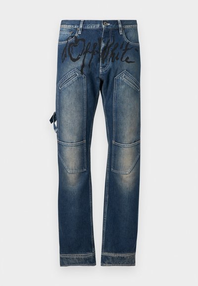 Pantalon en denim bleu avec des taches décolorées, plusieurs poches dont des poches avant inclinées, et un texte noir de style graffiti sur la partie supérieure avant.