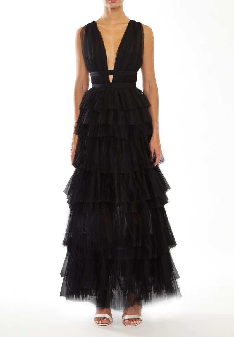 Femme vêtue d'une robe en tulle noire à étages sans manches avec un profond décolleté en V et des talons blancs ouverts à l'avant, debout sur un fond blanc.