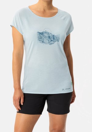 Kvinde iført lyseblå t-shirt med kort ærme med bjerggrafik og sorte shorts mod en ensfarvet baggrund.
