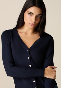 Jeune femme aux longs cheveux foncés portant un cardigan bleu marine boutonné, tenant son bras gauche avec un fond beige neutre.