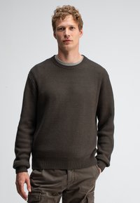 Dunkel gestreifter Strickpullover mit Rundhalsausschnitt, strukturiertem Oberflächenmuster und langen Ärmeln, kombiniert mit olivfarbenen Cargo-Hosen, Modell steht vor einem schlichten Hintergrund.