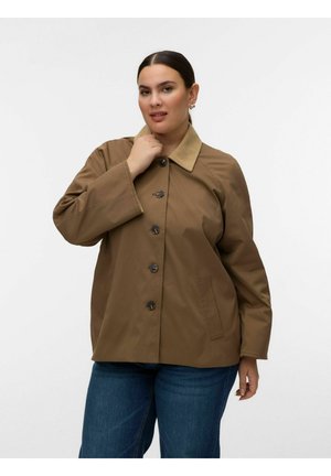 Femme aux cheveux foncés attachés en queue de cheval basse, portant une veste marron boutonnée avec un col beige et un jean bleu, sur un fond blanc uni.