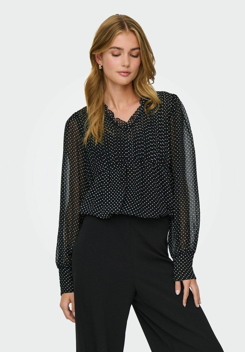 Femme portant un chemisier noir transparent à pois avec des manches longues et un pantalon noir taille haute, debout les yeux fermés.