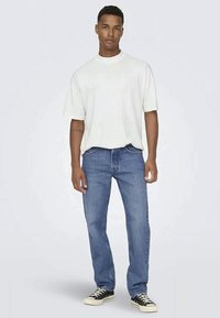 T-shirt branco de manga curta com gola redonda, combinado com jeans de denim azuis. O modelo usa sapatilhas pretas de cano baixo, em pé contra um fundo claro.