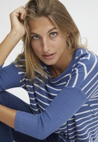 Blau gestreifter Pullover mit einem lockeren Ausschnitt und gerippten Bündchen, kombiniert mit dunkelblauen Jeans. Das Modell posiert mit einer Hand im Haar und zeigt ihre Gesichtszüge.