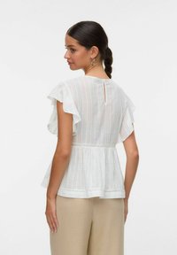 Witte peplumtop met fluttermouwen, voorzien van verticale streepjes, een keyhole achterkant en kantafwerking langs de zoom, gecombineerd met beige broek.