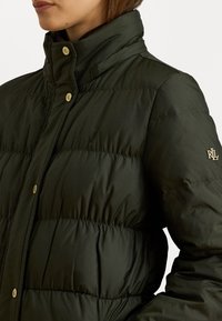 Olive zelená páperová bunda s vysokým golierom, vertikálnymi švami a zlatými kovovými doplnkami. Výrazný detail loga na rukáve.