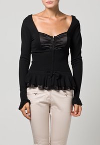 Nainen, jolla on päällään musta pitkähihaisista tiukka paita, jossa on vetoketjukiinnitys, poimutettu rintaosa, peplum-helma ja nauhadetaili. Housuina vaaleanbeiget housut, joissa on vetoketjutaskut.