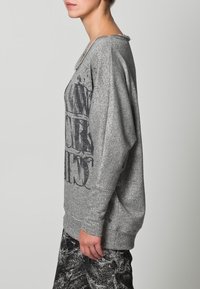 Srebrny, błyszczący sweter o luźnym kroju. Posiada okrągły dekolt i wyrazisty czarny napis na przedzie. Miękka, teksturowana tkanina.