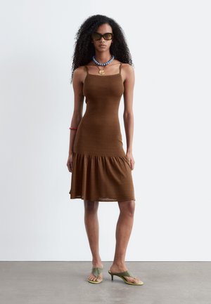 Vrouw met lang krullend haar, die een bruine mouwloze middellange jurk draagt, een blauwe kralenhalsketting, zonnebril en groene sandalen met hakken, binnen staand.