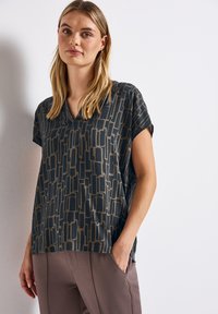 Top morbido e ampio di colore blu scuro con motivo geometrico beige, dotato di scollatura a V e maniche corte. Materiale soffice con un lieve lucido e caduta elegante.