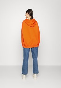Sudadera naranja de gran tamaño con cordón y bolsillo tipo canguro, combinada con jeans de denim azul acampanados y botas de plataforma blancas.