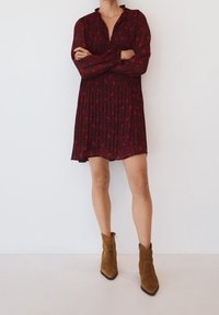 Punainen kukkakuosiset mekko, jossa on pleatattu muotoilu, pitkät hihat ja kauluksellinen kaulus. Yhdistetty ruskeisiin mokkajalkineisiin nilkkamittaisina.