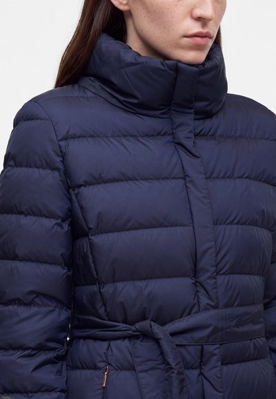 Femme portant une doudoune matelassée bleu marine avec un col montant et une ceinture nouée à la taille.