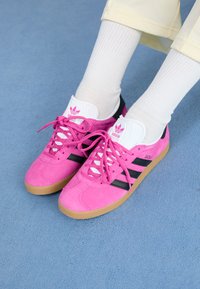 Pink suede sneakers with black accents and white laces. Features a white tongue with a logo, and a textured gum sole.