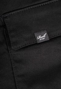 Close-up of black fabric pocket with stitched flap and sewn-in label reading "Reell est. 1997" in white script.
