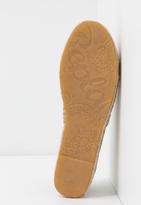 Semelle d'une chaussure en caoutchouc beige avec le nom de la marque et des motifs décoratifs embossés sur la semelle, positionnée droite contre un coin blanc.