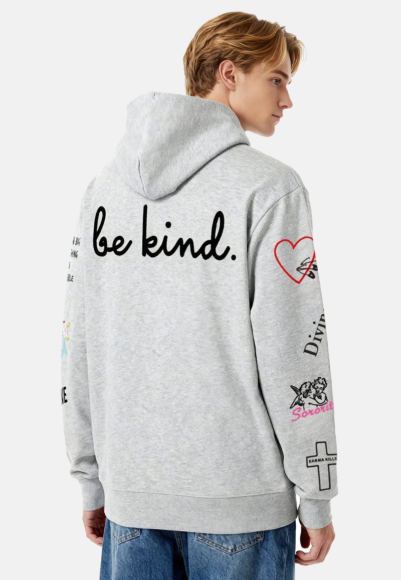 Harmaa hupullinen svetari, jossa teksti "be kind.", koristeellisia kuvioita hihoissa ja erilaisia painokuvioita, yksinkertaisella taustalla.