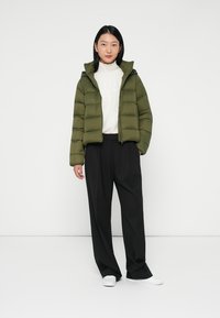 Zeleno-olivová péřová bunda s kapucí, prošívaný design, přední zip, nošena přes bílý pletený svetr a černé široké kalhoty.