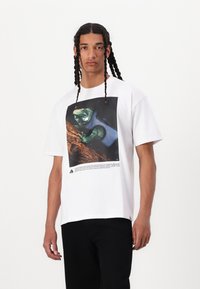 Tricou alb cu mânecă scurtă, prezentând un grafic viu al unei șopârle pe un fundal texturat, cu text imprimat sub imagine.