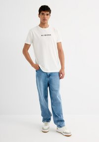 Camiseta de algodón blanca con texto impreso en negro "TODO LO QUE VEO ES AZUL", combinada con jeans de un azul claro y zapatillas blancas. Diseño simple y casual.