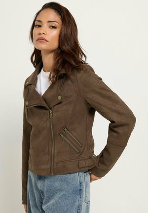 Femme aux cheveux bruns mi-longs portant une veste brune zippée sur une chemise blanche et un jean bleu clair, debout, main sur la hanche.