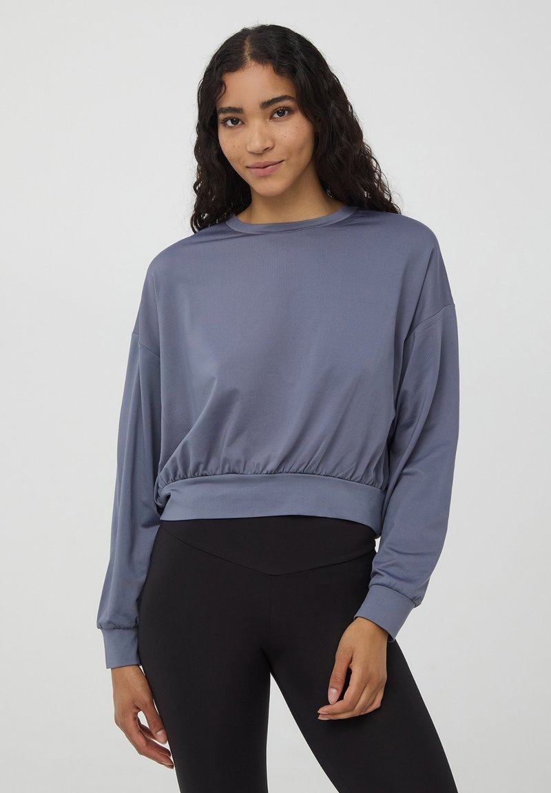 Fioletowy, krótki sweatshirt z długimi rękawami, okrągłym dekoltem i marszczonym dołem. Gładka tkanina o subtelnym połysku, zestawiona z czarnymi legginsami.