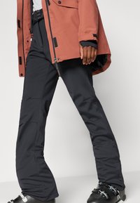 Παντελόνια σκι σε ματ μαύρο χρώμα, με στενή γραμμή και διακριτικές ραφές. Συνδυάζονται με ένα μπουφάν σκουριάς, με πολλές τσέπες και φερμουάρ.