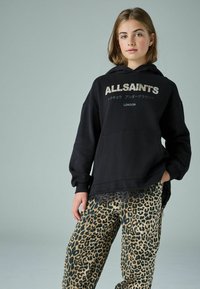 Čierna mikina s kapucňou a logom "ALLSAINT'S" v metalickom písme, v kombinácii so zelenými nohavicami s leopardím vzorom, ktoré majú voľný strih a vrecká.
