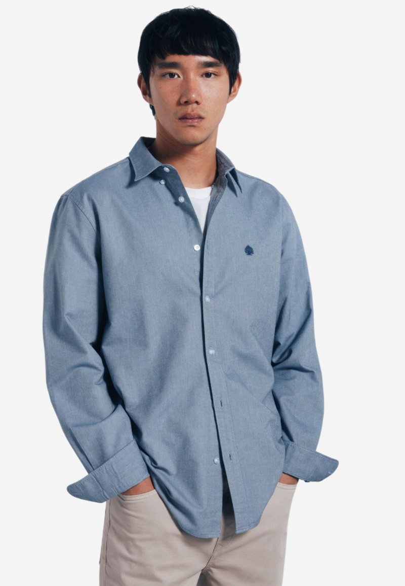 Camicia blu chiaro con bottoni, caratterizzata da un motivo a quadretti discreto, una texture morbida e un piccolo logo ricamato sul petto. Indossata sopra una maglietta bianca.