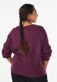 Gruby purpurowy sweter z prążkowanymi mankietami i dolnym obszyciem, w luźnym kroju i teksturowanym wzorze dzianiny. Włosy modelki zaplecione w warkocz z tyłu.