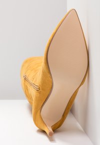 Botte stiletto en daim jaune avec un bout pointu et une tige texturée. Dotée d'un détail zippé sur le côté et d'une semelle lisse et claire.