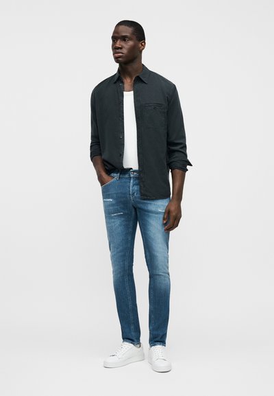 Chemise boutonnée vert foncé portée sur un débardeur blanc, associée à un jean slim bleu clair légèrement usé et des baskets blanches.