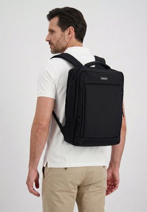 Man in wit poloshirt en beige broek draagt een zwarte rechthoekige rugzak met het opschrift "Venezia" tegen een effen lichte achtergrond.