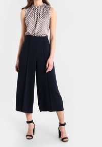 Blouse à pois beige et blanc avec col montant ; pantalons larges bleu marine ; sandales noires à talons carrés. Tissu lisse avec drapé.