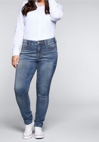 Modré džíny skinny střihu se středním vypráním, s pěti kapsami a kovovým knoflíkovým zapínáním, v kombinaci s bílou košilí na knoflíky a bílými teniskami.