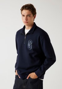 Marineblauwe sweatshirt met een kwart-rits, gemaakt van textuurstof, met ribboord manchetten en geborduurd logo op de borst. Gecombineerd met spijkerbroek.