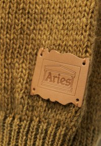 Καφέ πλεκτό ύφασμα με ραμμένη δερμάτινη ετικέτα που γράφει «Aries» και αρχιτεκτονικό σχέδιο κολώνας πάνω από το κείμενο.