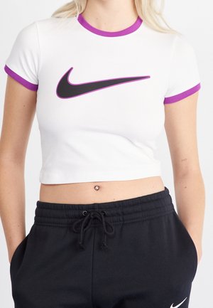 Kobieta nosząca białą krótka koszulkę z fioletowym obszyciem i czarnym logo Nike, w połączeniu z czarnymi spodniami ze ściągaczem i kieszeniami.