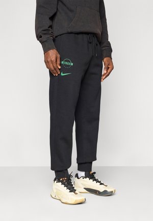 Homme portant un pantalon de survêtement noir des Celtics et des baskets beige, se tenant contre un fond clair uni.