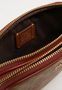 Sac en cuir marron avec fermeture éclair dorée, intérieur doublé de tissu marron. Comprend une étiquette de marque beige et un extérieur texturé.