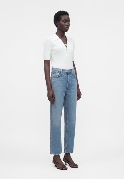 Γυναίκα με λευκό ριπ τοπ με κοντά μανίκια, ανοιχτό μπλε τζιν με ψηλή μέση και ίσια γραμμή, και μαύρα σανδάλια με χαμηλό τακούνι, στέκεται μπροστά σε απλό φόντο.
