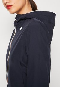 Granatowa kurtka z kapturem zapinana na zamek błyskawiczny, z kontrastowym wykończeniem zamka i detalem z logo na piersi. Gładki, lekki materiał.